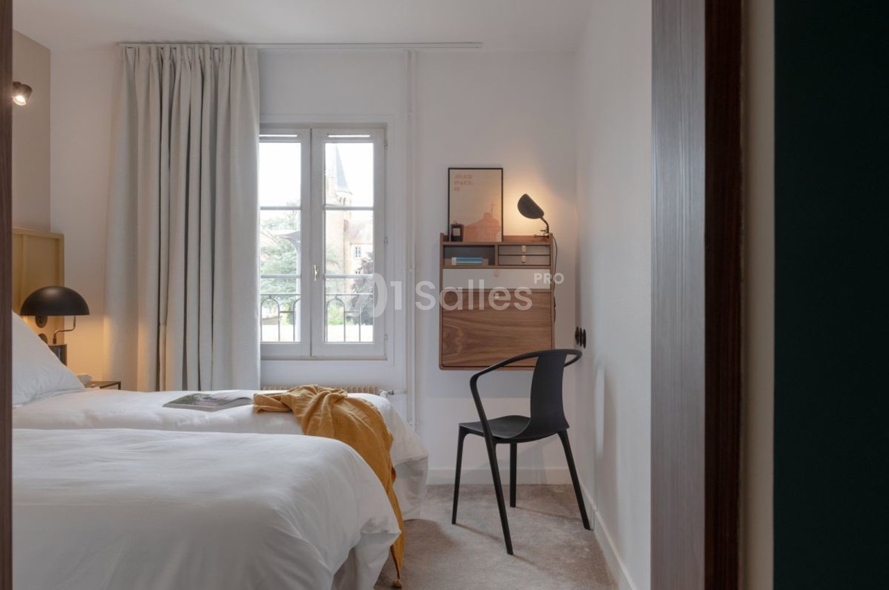 Chambre lumineuse avec lit double, bureau mural en bois, chaise noire et fenêtre donnant sur un paysage extérieur.