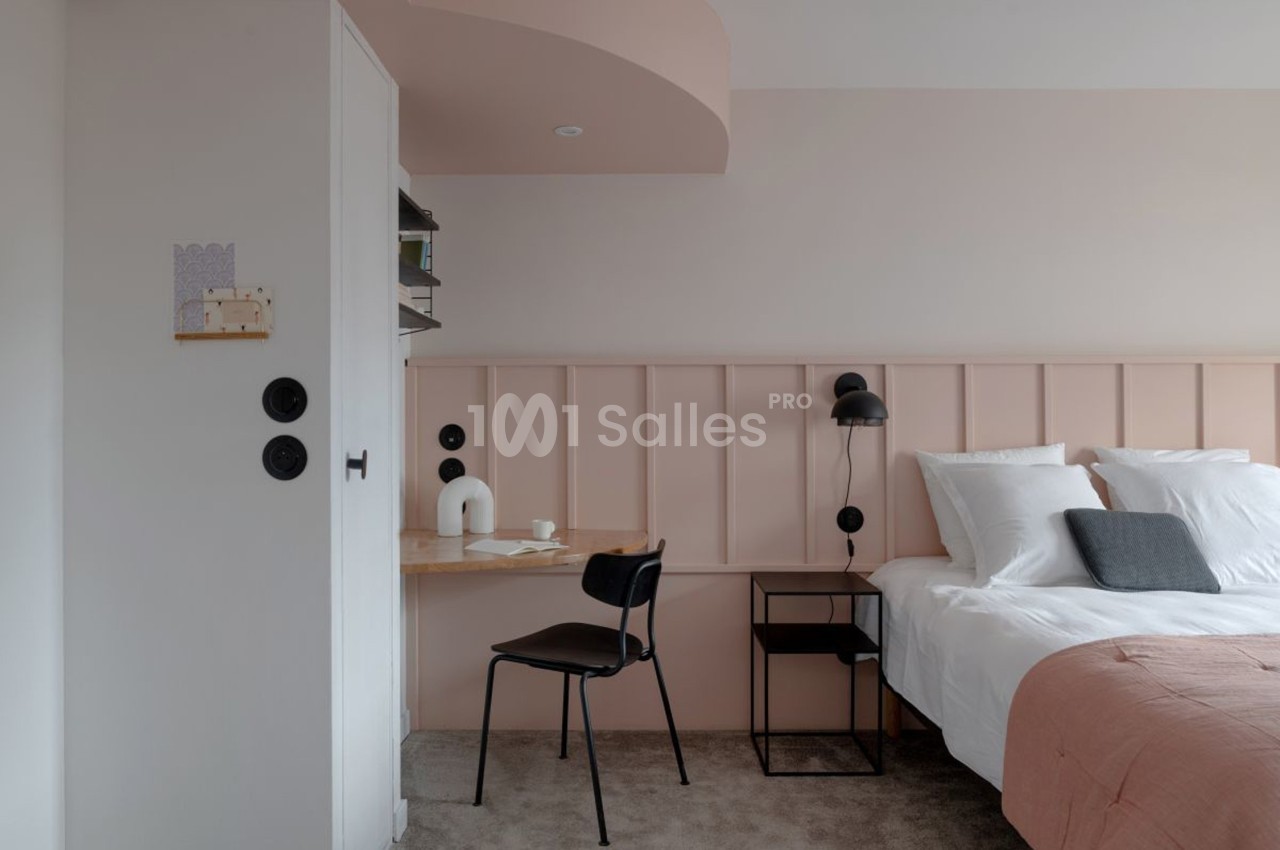 Chambre moderne avec lit double, mur rose, bureau en bois clair, chaise noire et éclairage mural.