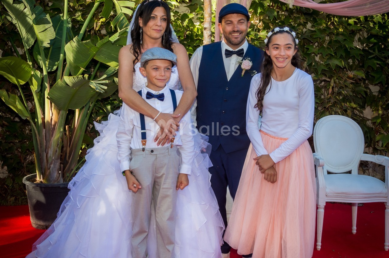 Un couple en tenue de mariage pose avec deux enfants sur un tapis rouge, devant un décor végétal et floral.