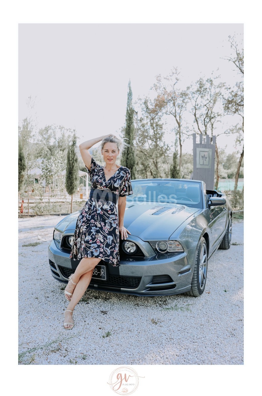 Femme en robe fleurie posant devant une voiture décapotable grise, dans un environnement extérieur arboré.