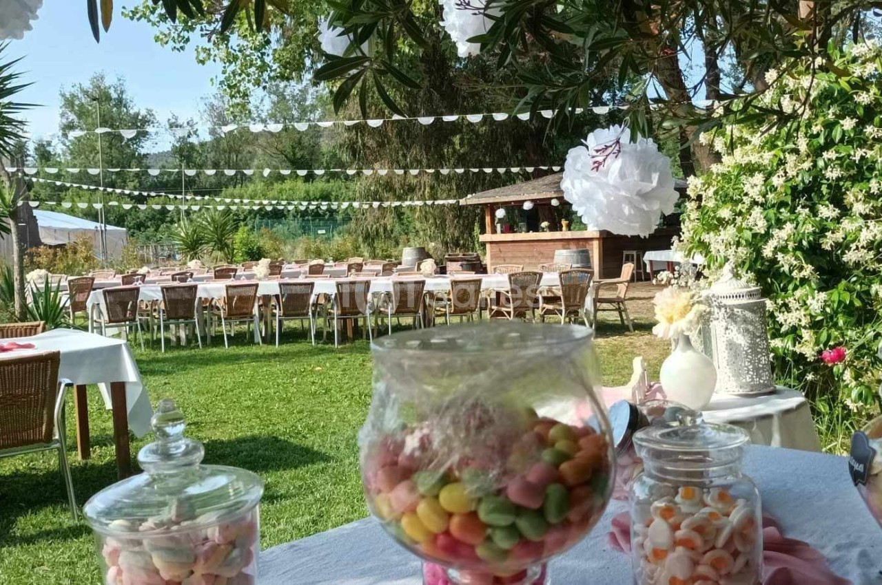 Tables dressées en extérieur sous des guirlandes, avec des bonbonnières au premier plan et un jardin fleuri en arrière-plan.