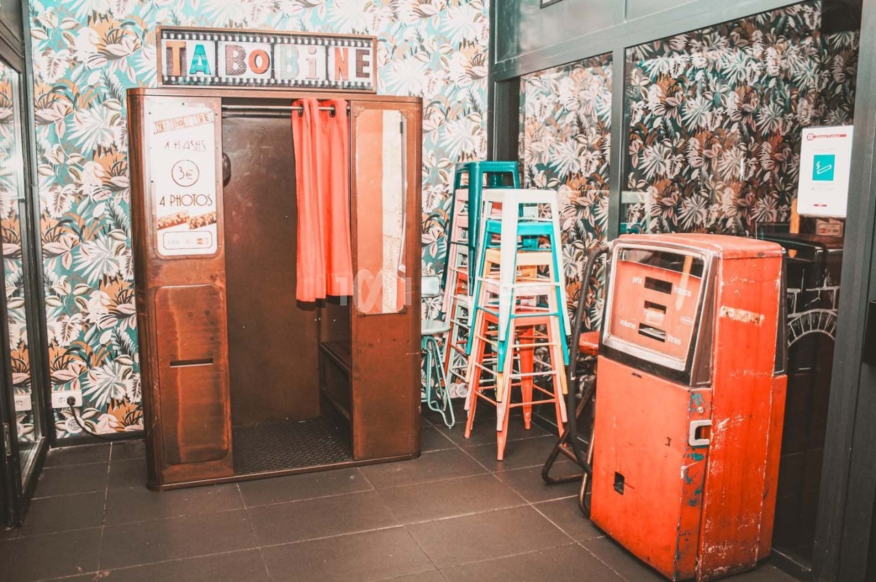 Cabine photo vintage avec rideaux rouges, entourée de tabourets empilés et d'un distributeur automatique rouge.
