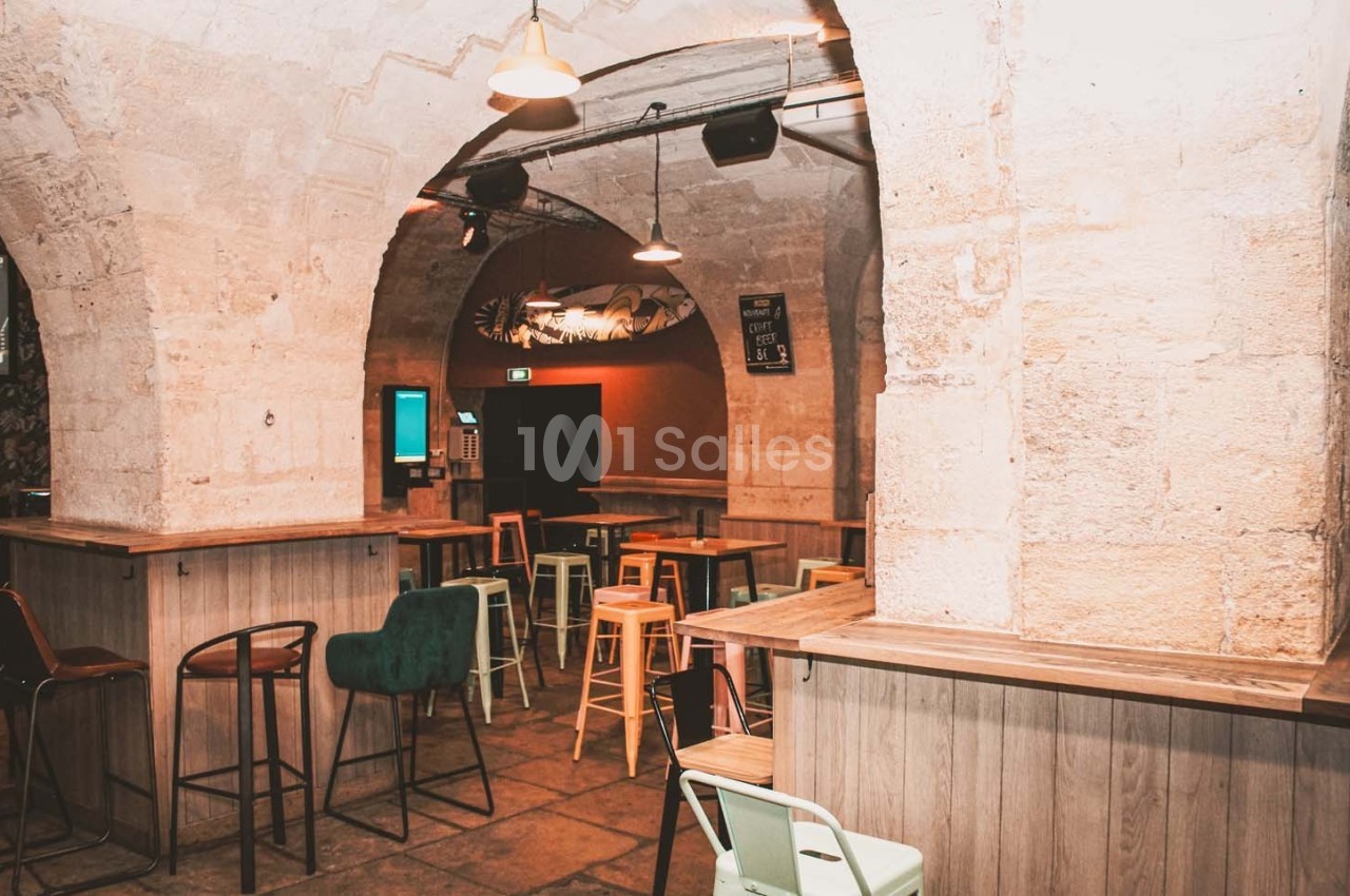 Intérieur d'un bar avec des voûtes en pierre, des tables hautes et des chaises colorées dans une ambiance chaleureuse.