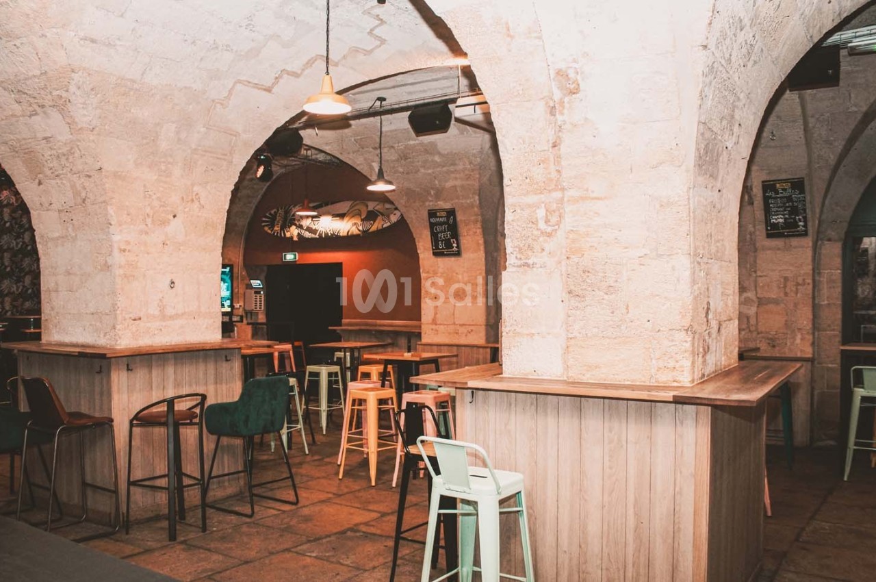 Salle voûtée en pierre avec des tables hautes, des tabourets colorés et un éclairage tamisé.