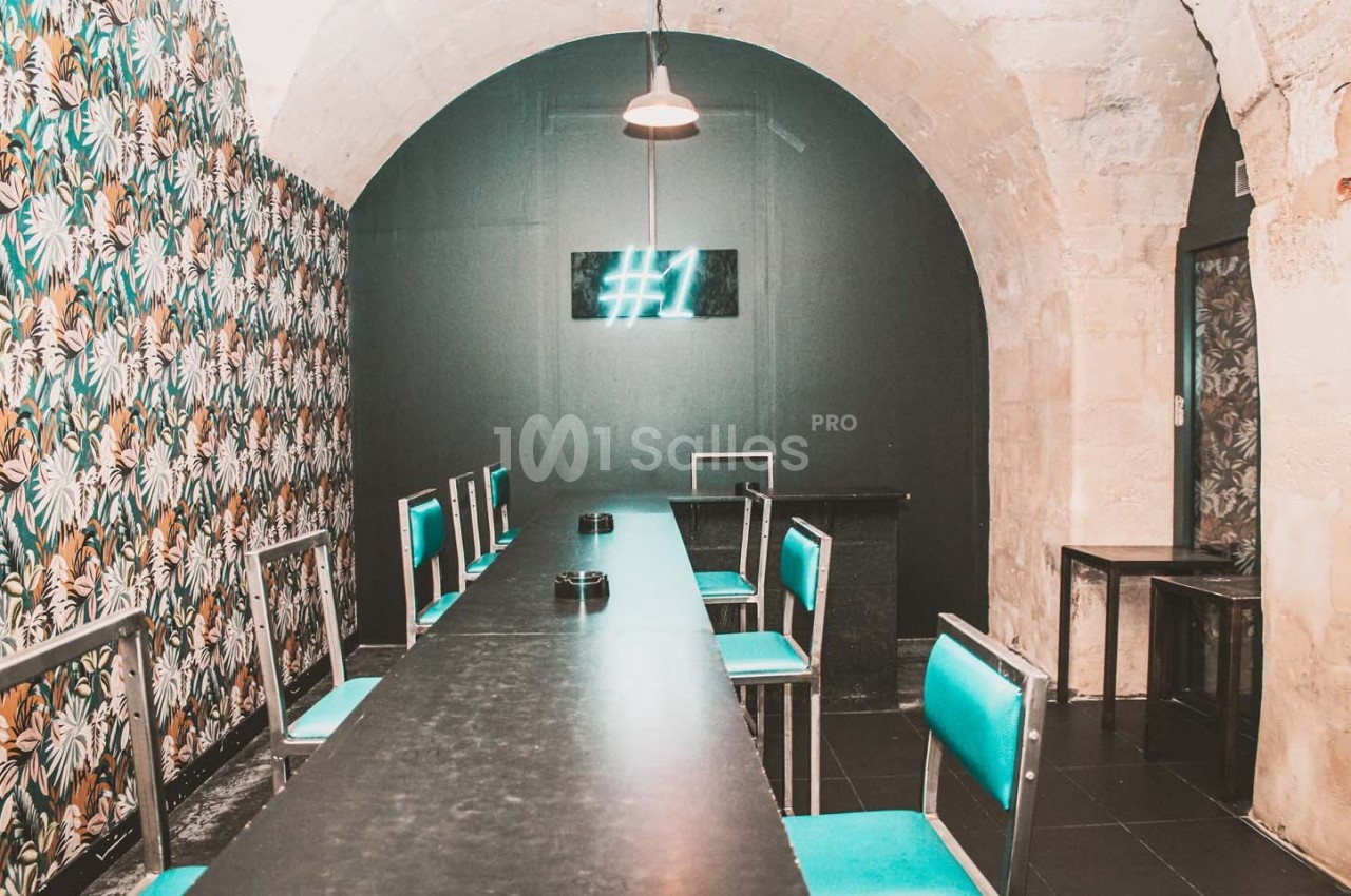 Salle voûtée avec une longue table noire, chaises turquoise, mur fleuri et enseigne lumineuse ’#1’.