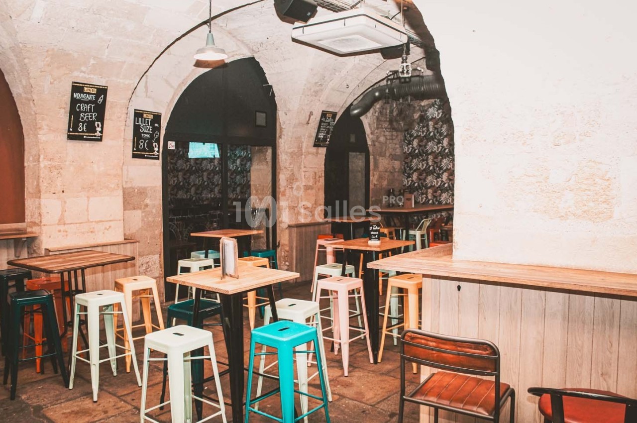 Salle de bar avec tables hautes, tabourets colorés et murs en pierre voûtés.