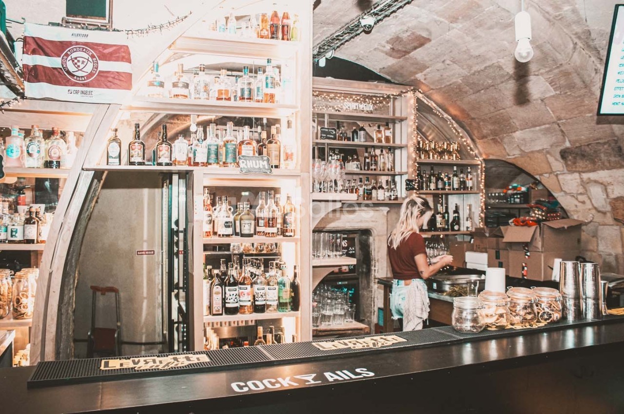 Bar voûté avec étagères remplies de bouteilles, une personne prépare des boissons derrière le comptoir.