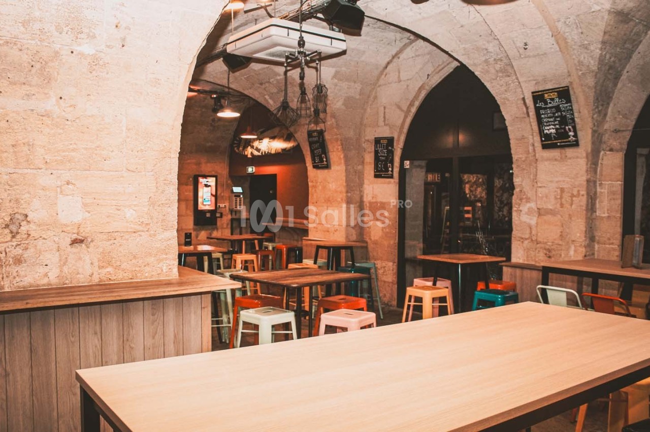 Salle voûtée avec murs en pierre, tables en bois et tabourets colorés, éclairée par des suspensions industrielles.