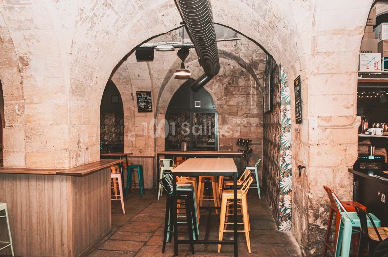 Salle voûtée en pierre avec tables hautes, tabourets colorés et décoration lumineuse dans un cadre chaleureux.