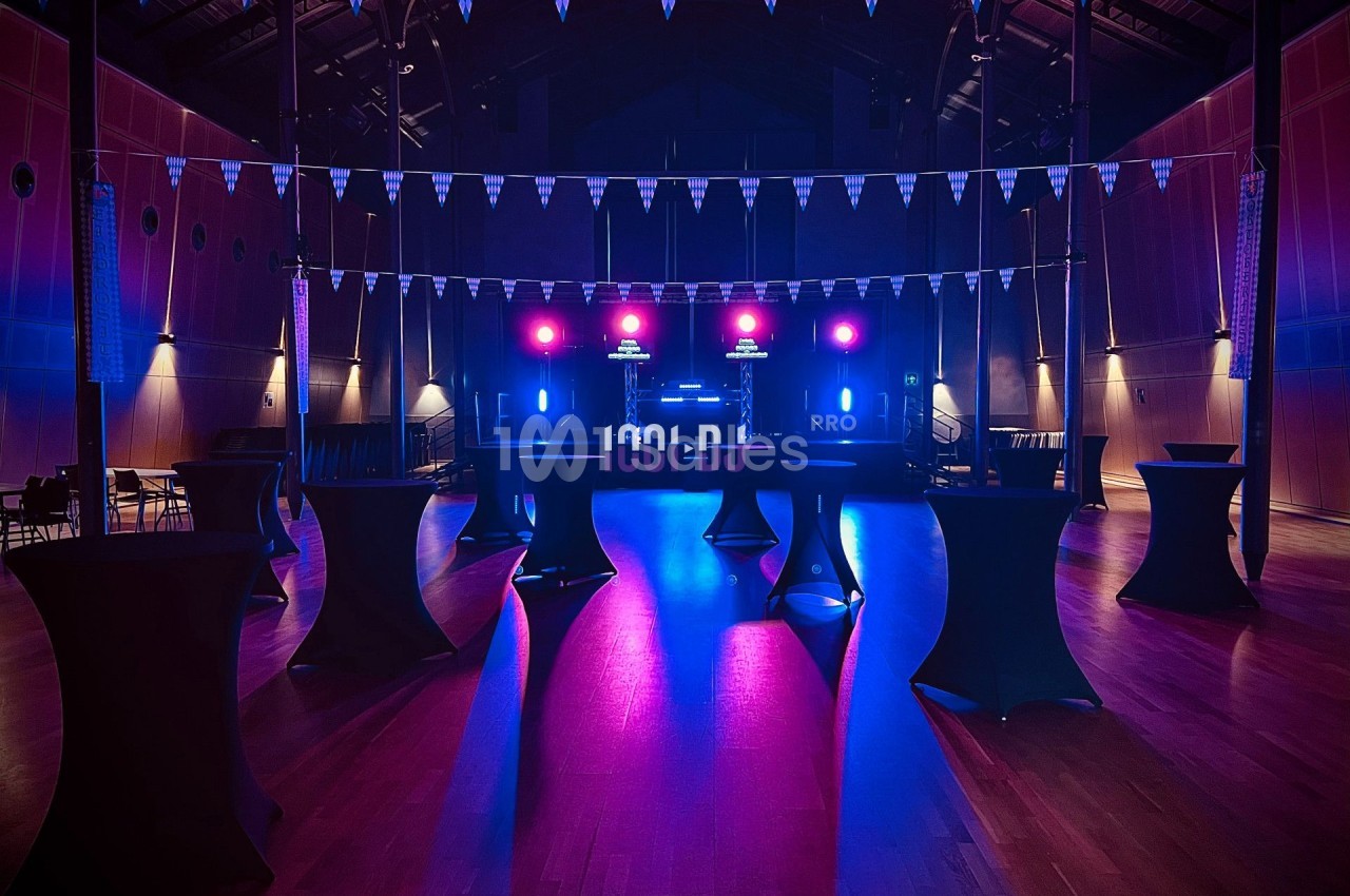 Salle de réception avec éclairage coloré, tables hautes noires et scène équipée de lumières et matériel audio.