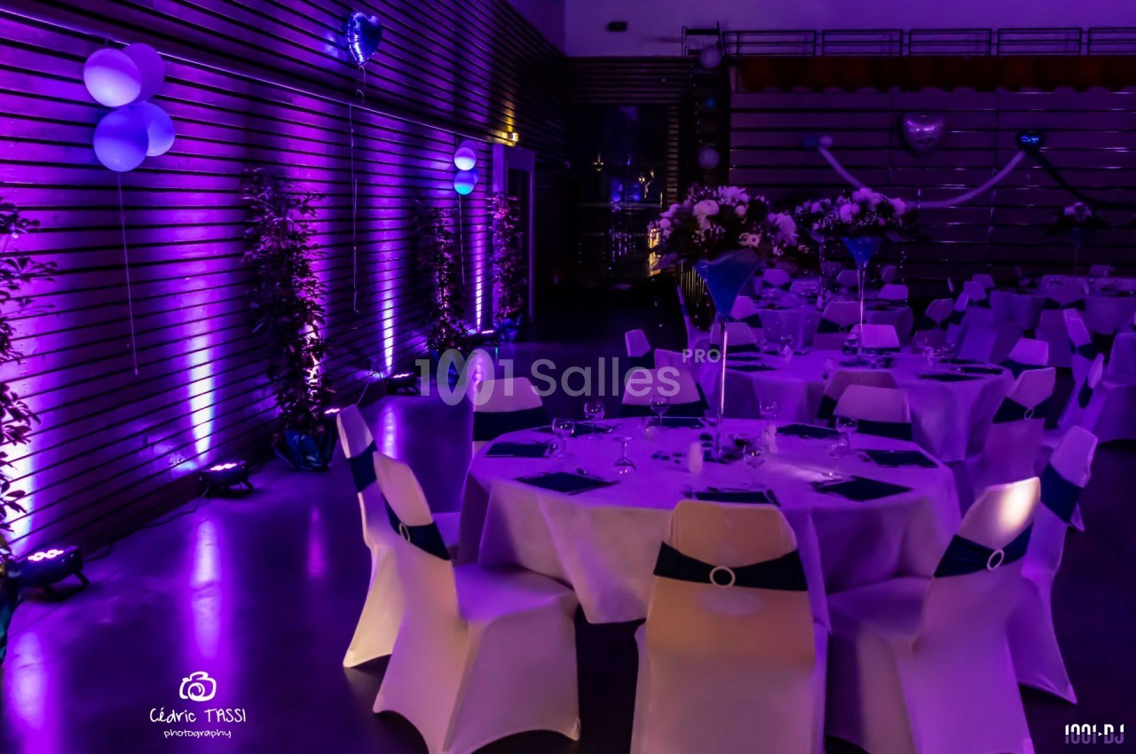 Salle décorée pour un événement, avec des tables rondes dressées et un éclairage violet créant une ambiance festive.