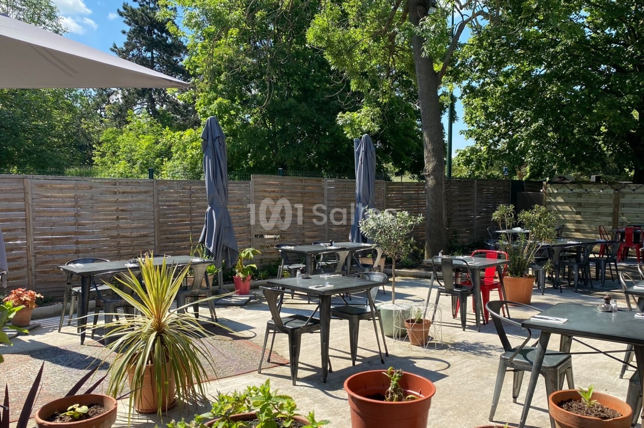 Terrasse extérieure avec tables, chaises, parasols et plantes en pots, entourée de végétation et d'une clôture en bois.