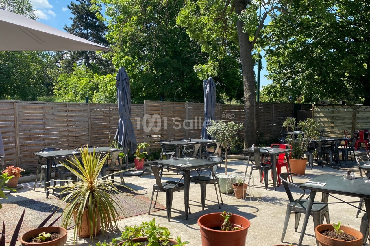 Terrasse extérieure avec tables, chaises, parasols et plantes en pots, entourée de verdure et d'une clôture en bois.