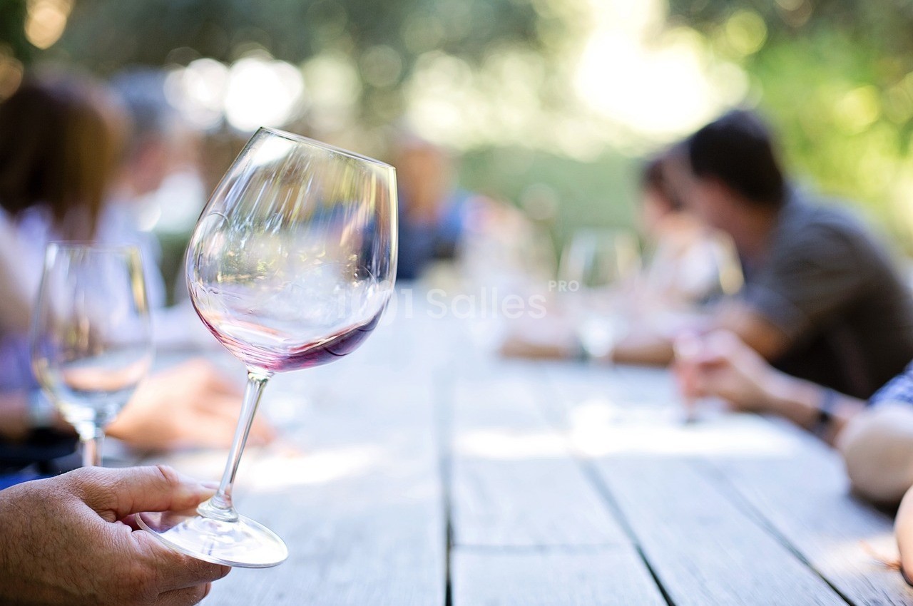 Un verre de vin rouge tenu à la main sur une table en bois, avec des personnes floues en arrière-plan.