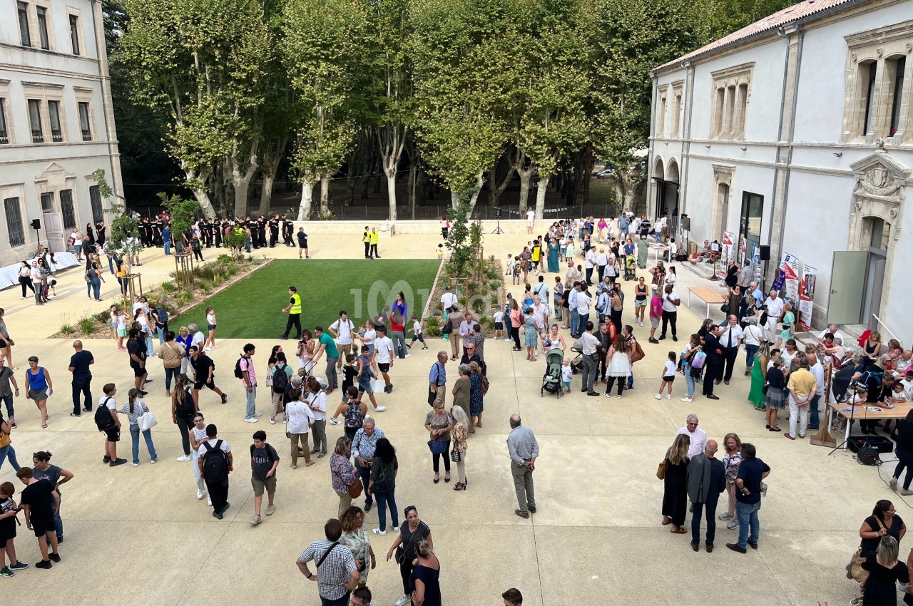 Foule rassemblée dans une cour extérieure bordée d'arbres et de bâtiments historiques par une journée ensoleillée.