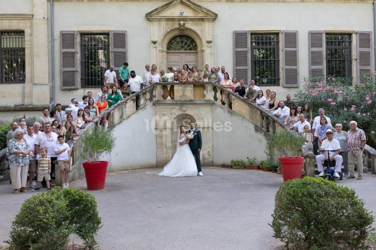 Un couple de mariés pose sur un escalier entouré de nombreux invités devant un bâtiment ancien.