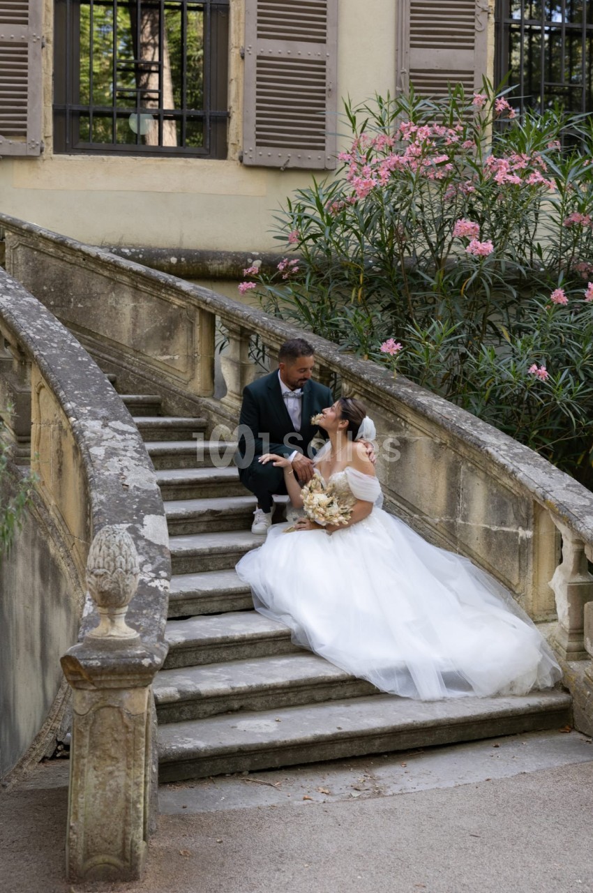 Un couple de mariés assis sur un escalier en pierre, entouré de fleurs et d'une végétation luxuriante.