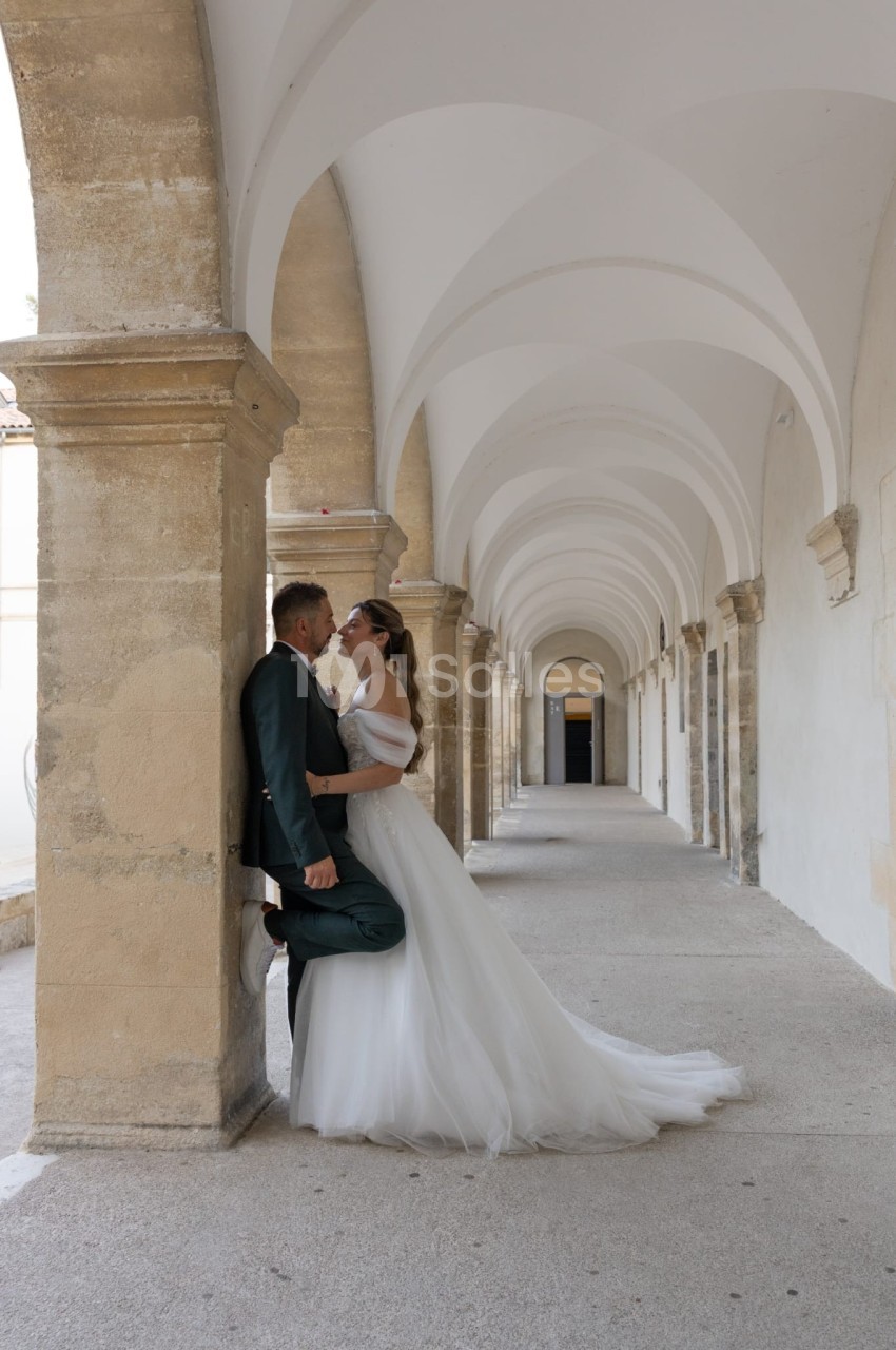 Un couple en tenue de mariage pose sous une galerie voûtée en pierre, dans un décor lumineux et élégant.