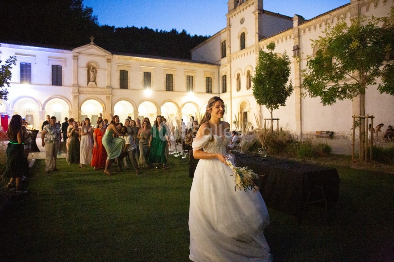 Une mariée en robe blanche s'apprête à lancer son bouquet devant un groupe d'invités dans une cour éclairée en soirée.