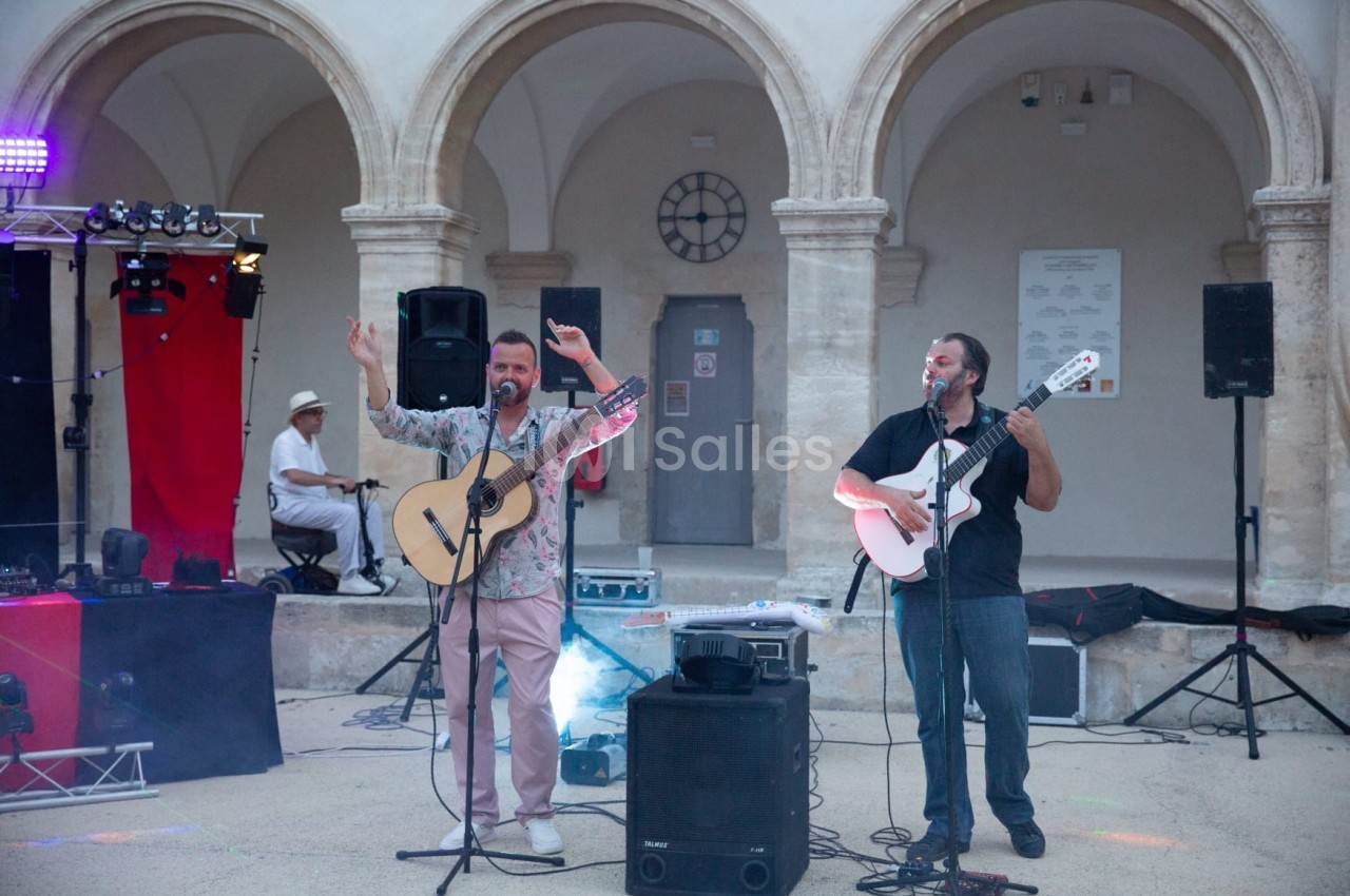 Deux musiciens jouent de la guitare et chantent sur une scène en plein air, devant des arches en pierre.