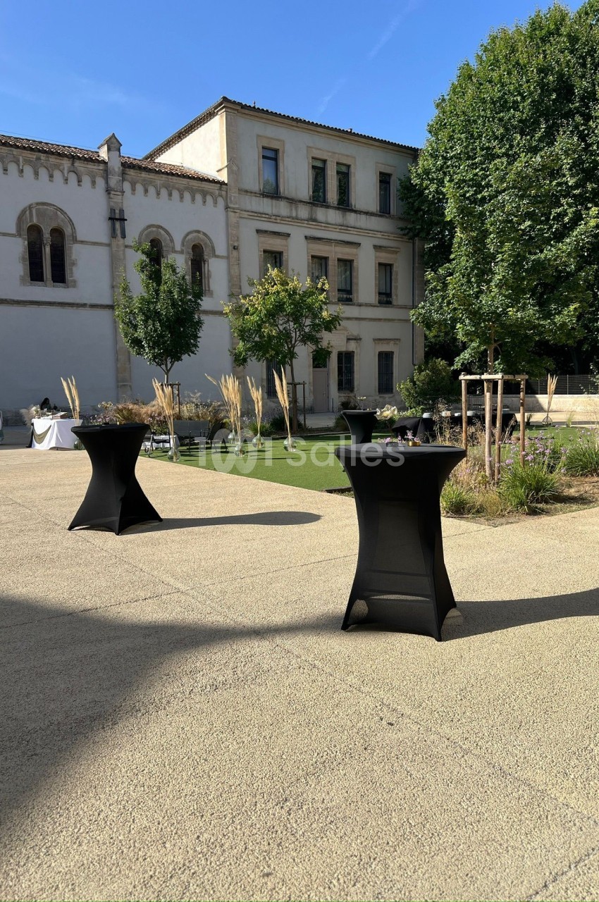 Cour extérieure ensoleillée avec des tables hautes noires, des plantes et un bâtiment ancien en arrière-plan.