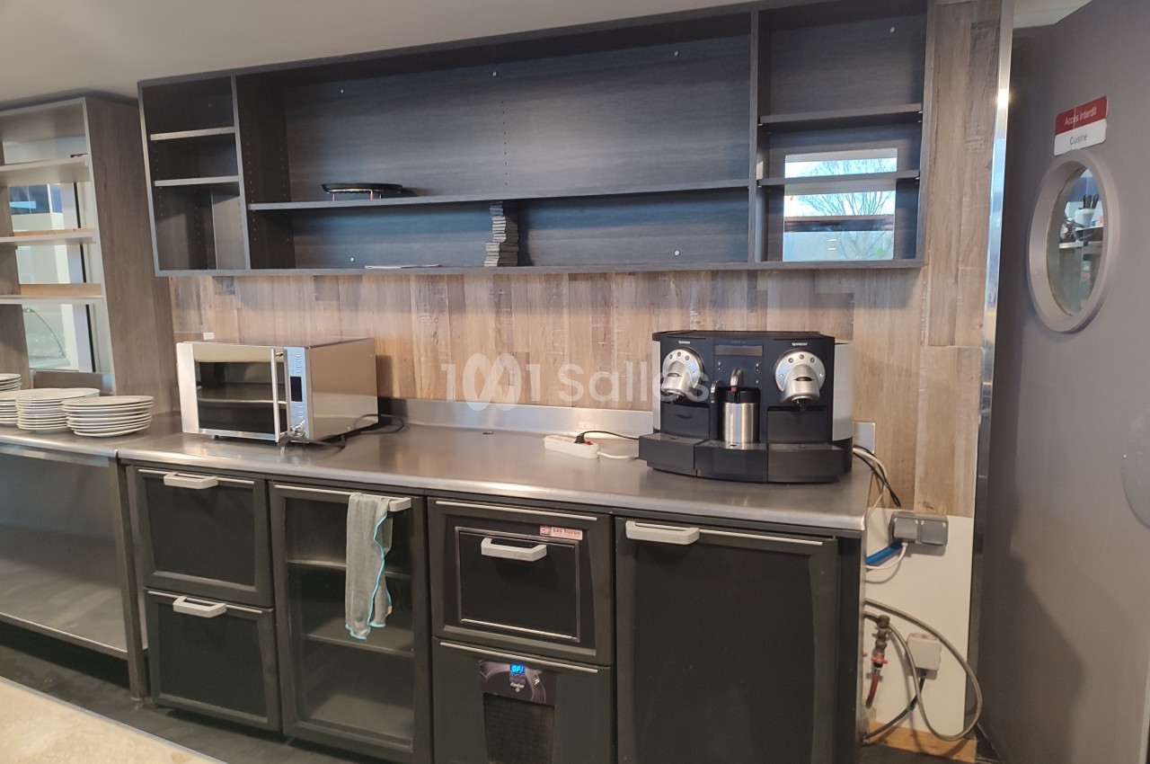 Cuisine équipée avec étagères vides, micro-ondes, machine à café et rangements noirs sous un plan de travail en inox.