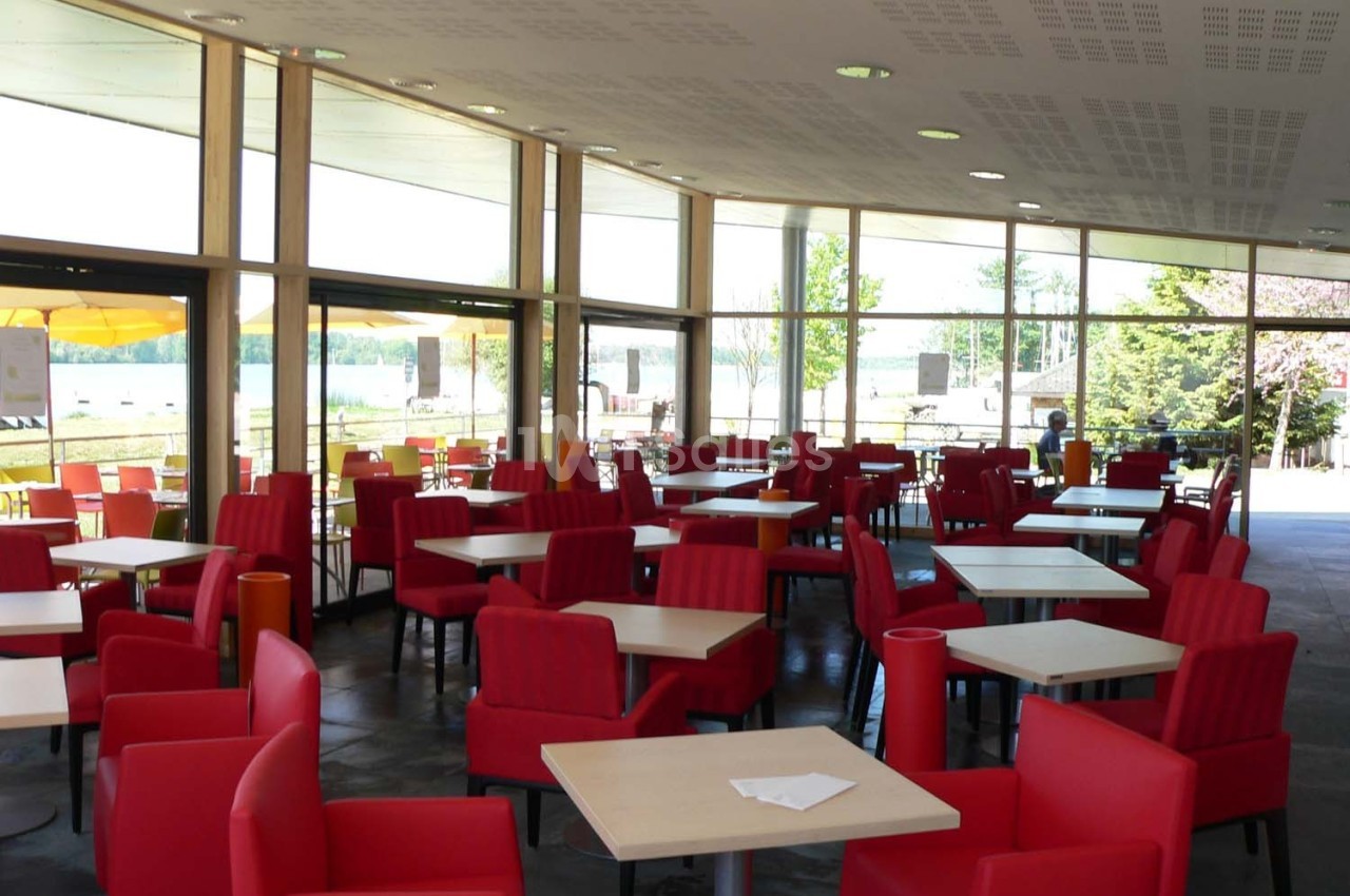 Salle lumineuse avec de grandes baies vitrées, des tables carrées et des fauteuils rouges disposés de manière espacée.