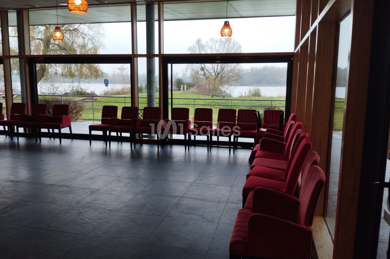 Salle lumineuse avec des chaises rouges alignées près de grandes baies vitrées donnant sur un paysage extérieur verdoyant.