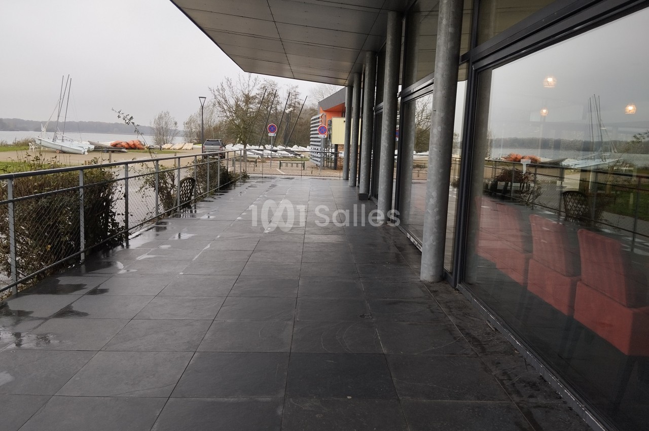 Terrasse couverte avec sol en dalles sombres, vue sur un lac, des voiliers et des arbres par temps nuageux.