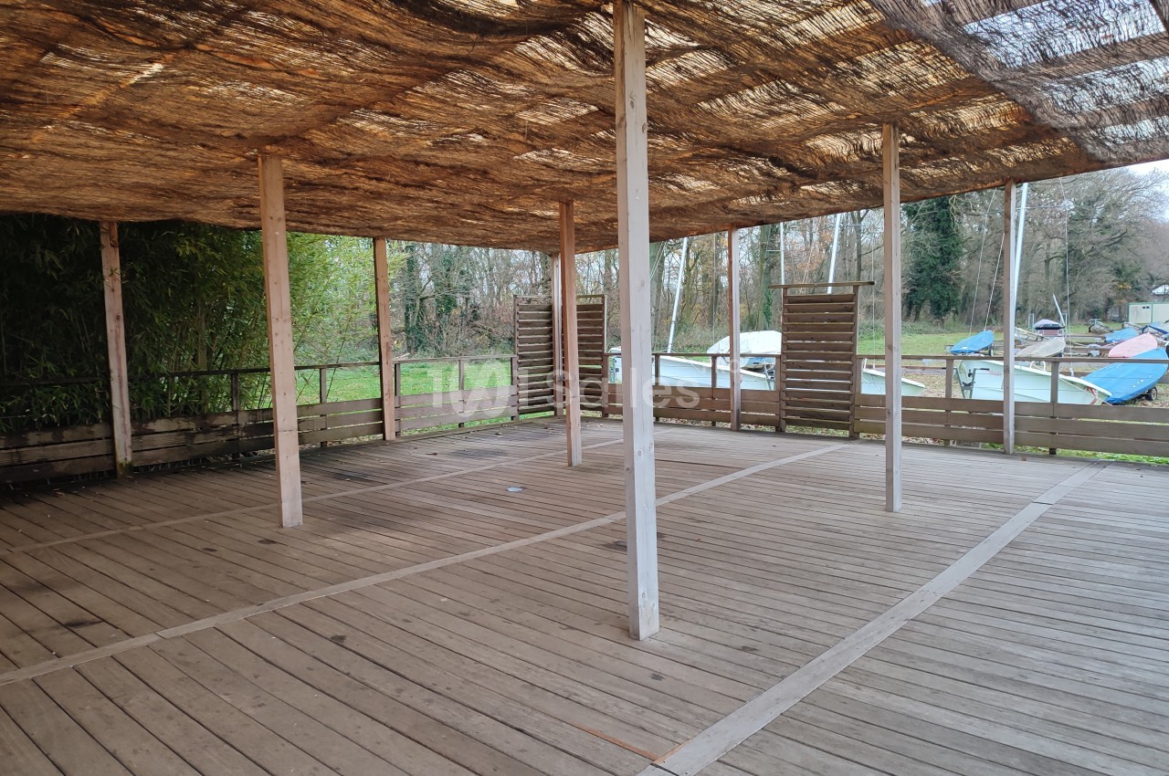Structure en bois avec toit en canisses, ouverte sur les côtés, située près d'un espace naturel avec des bateaux visibles.