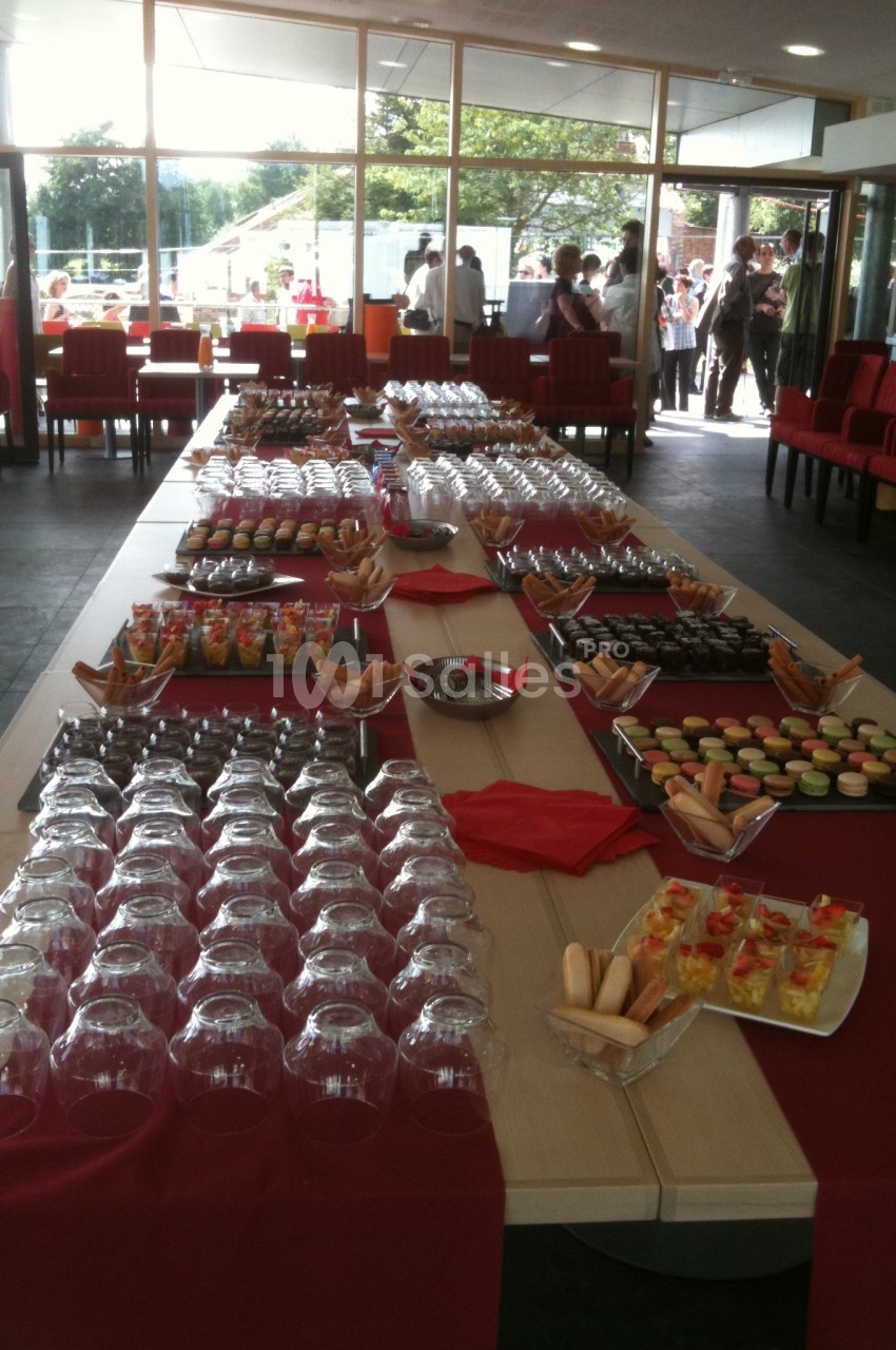 Table dressée avec des verrines, macarons, biscuits et verres, dans une salle lumineuse avec des invités en arrière-plan.
