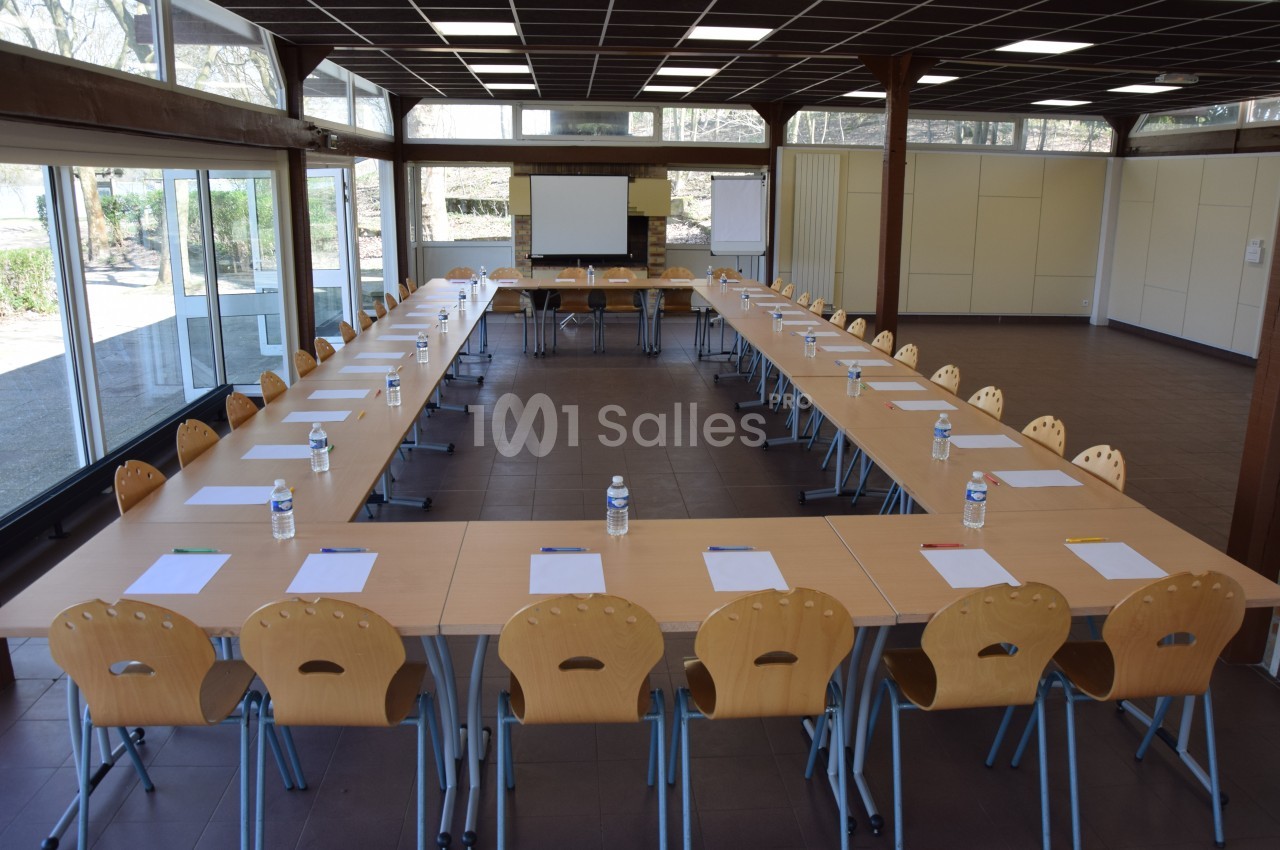 Salle de réunion lumineuse avec tables disposées en U, chaises en bois, paperboards et écran de projection.