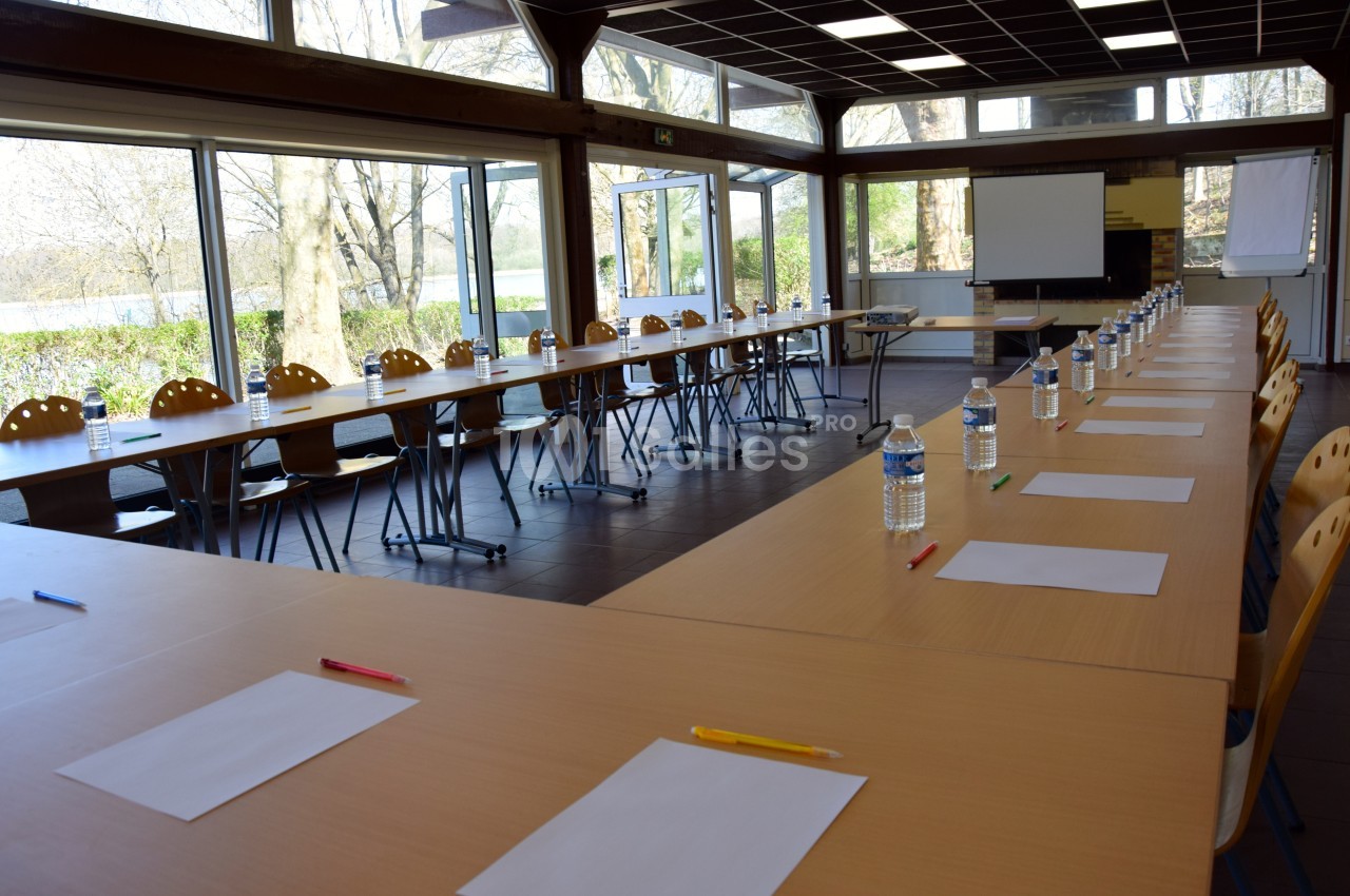 Salle de réunion lumineuse avec tables disposées en U, feuilles, stylos et bouteilles d'eau alignés pour chaque place.