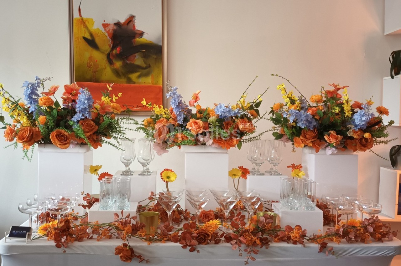 Table décorée avec des fleurs orange, jaunes et bleues, entourant des verres en cristal, dans un cadre élégant.