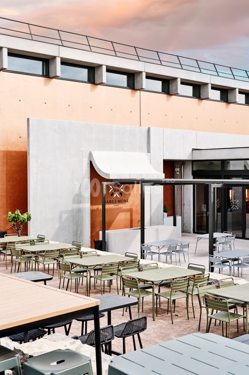 Terrasse extérieure avec tables et chaises modernes devant un bâtiment en béton et façade orange sous un ciel coloré.