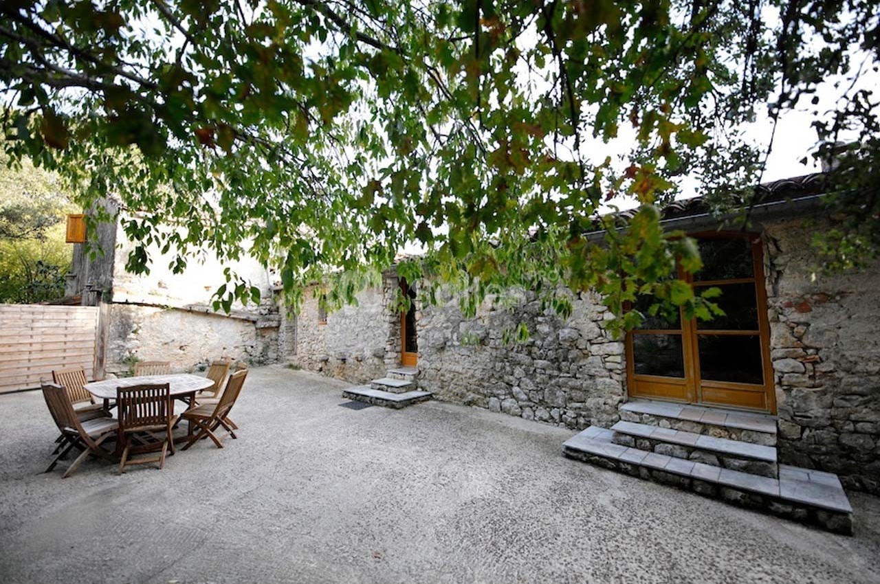Cour extérieure avec table et chaises en bois, entourée de murs en pierre et partiellement ombragée par des arbres.
