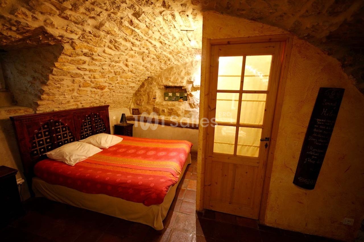 Chambre avec murs en pierre apparente, lit double avec couvre-lit rouge et porte vitrée donnant sur une pièce éclairée.
