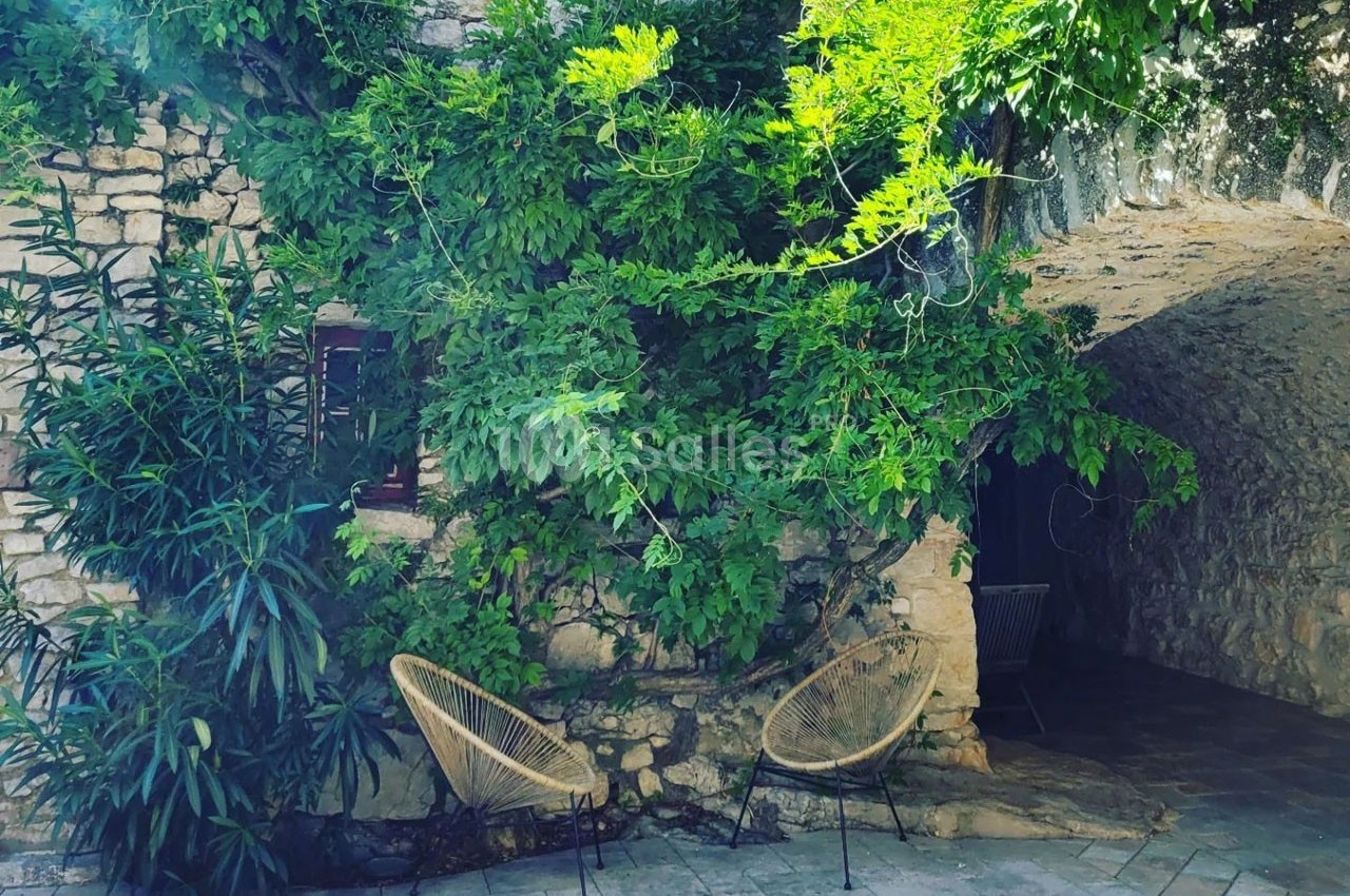 Mur en pierre recouvert de verdure avec deux fauteuils en osier disposés sur une terrasse pavée.