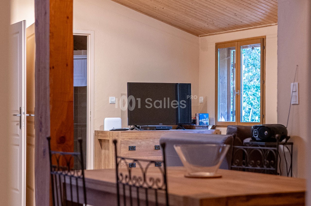 Intérieur d'une pièce avec table en bois, télévision sur un meuble, fenêtre donnant sur un extérieur arboré.