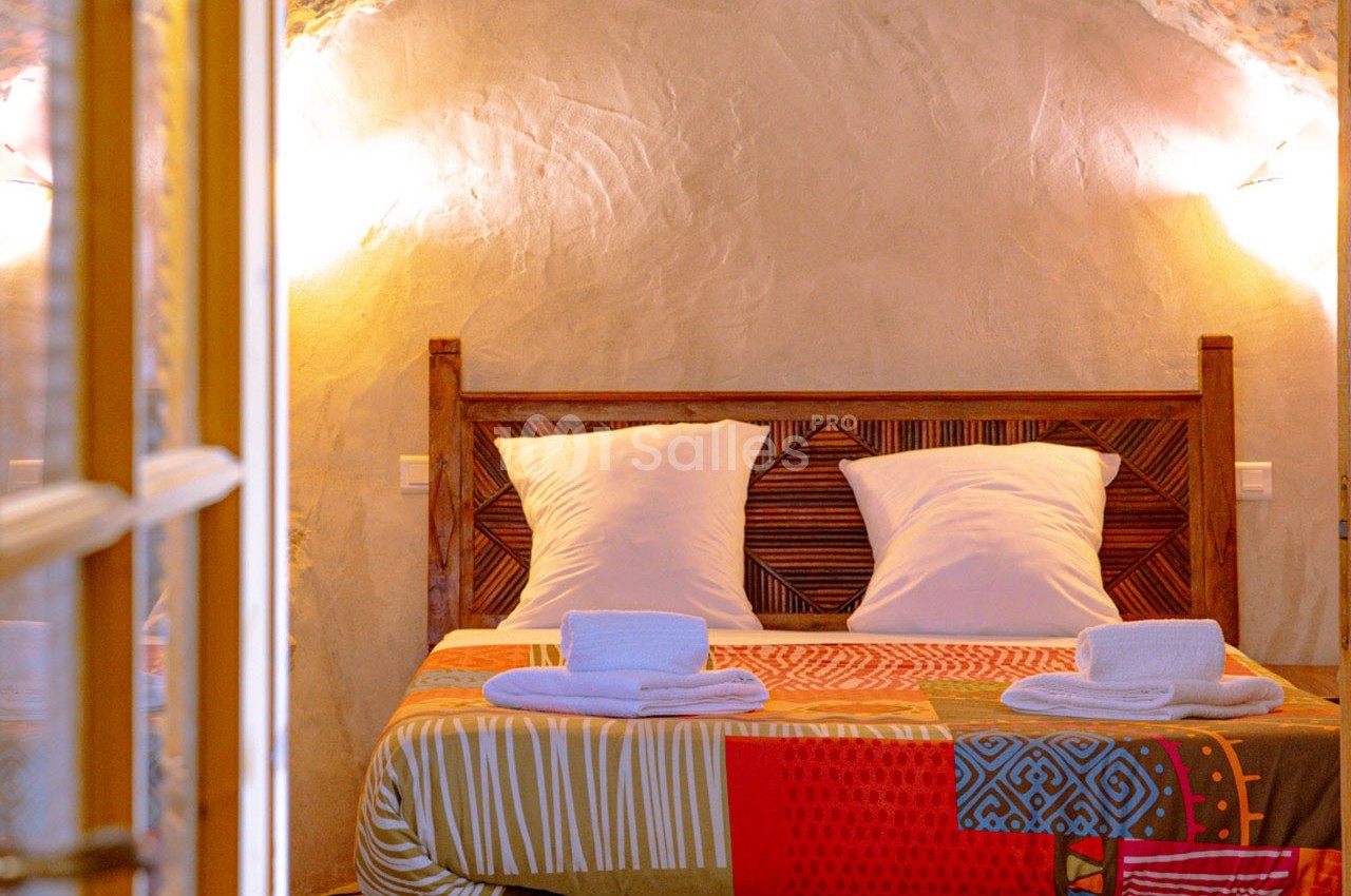 Lit double avec tête de lit en bois sculpté, draps colorés et serviettes pliées, dans une chambre aux murs en pierre.