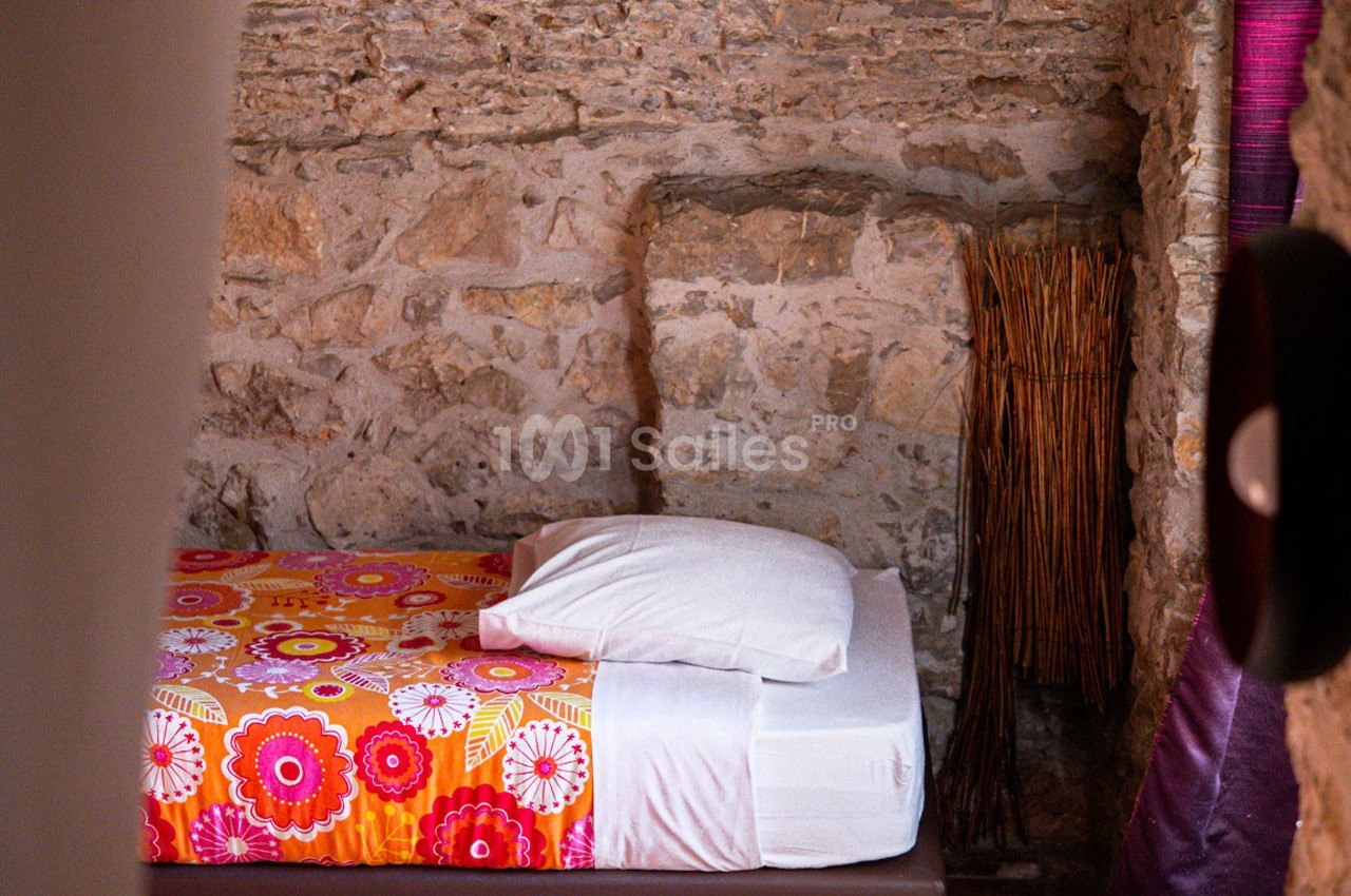 Lit simple avec une housse colorée à motifs floraux, adossé à un mur en pierre apparente dans une pièce rustique.