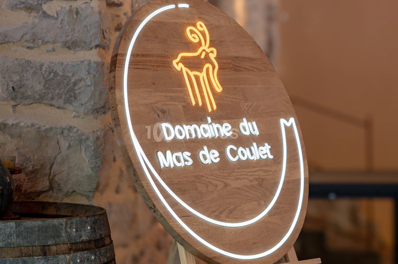 Panneau en bois circulaire avec logo lumineux et texte ’Domaine du Mas de Coulet’, posé sur un chevalet en intérieur.