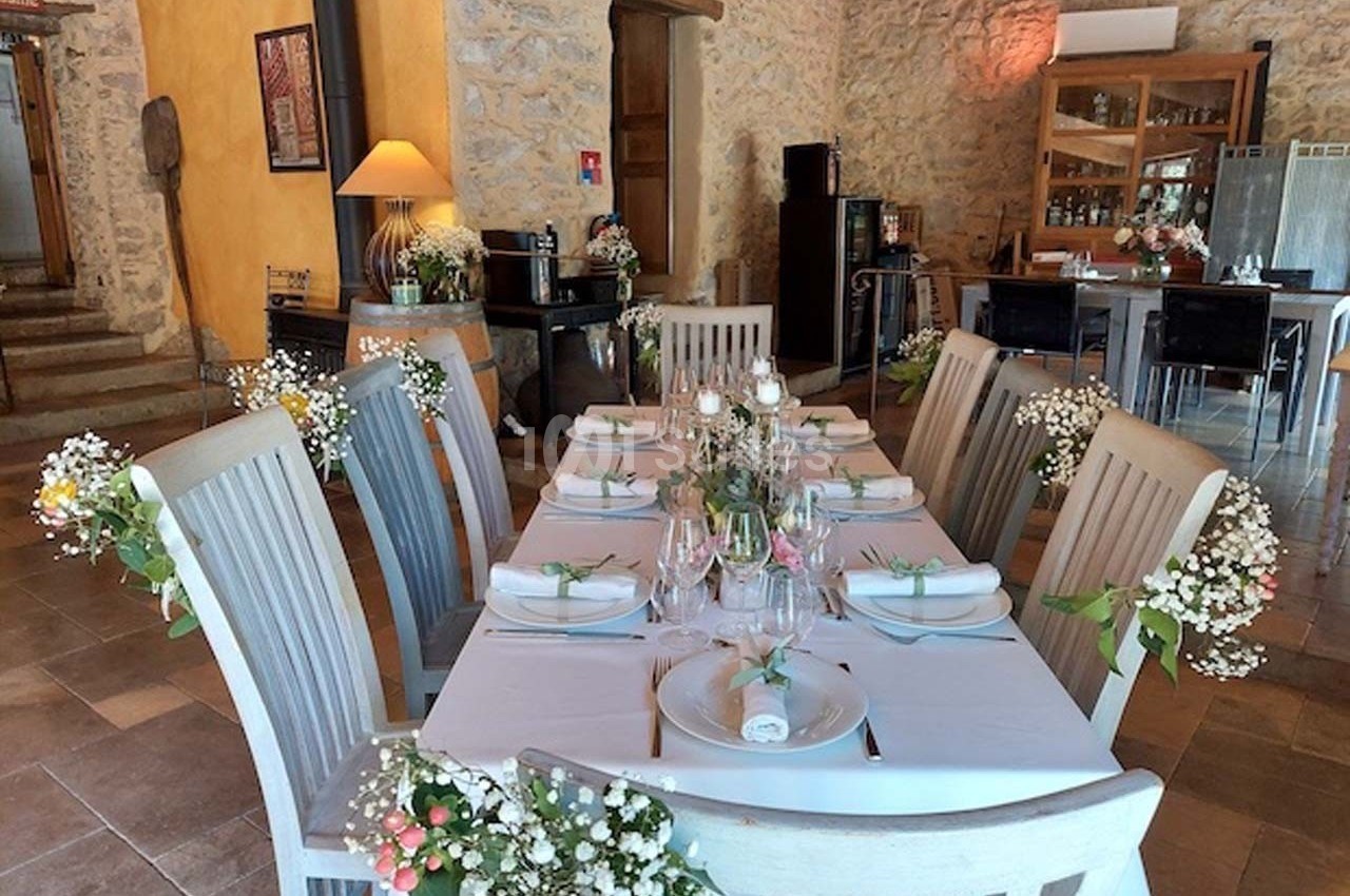 Table décorée pour un repas, avec nappes blanches, fleurs et chaises en bois, dans une salle en pierre lumineuse.
