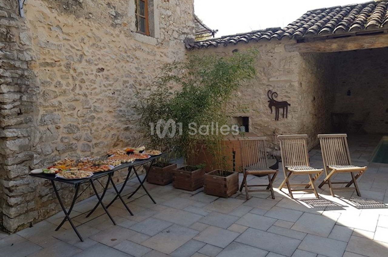 Cour en pierre avec table garnie de plats, chaises en bois alignées et mur décoré d'une peinture de chèvre.