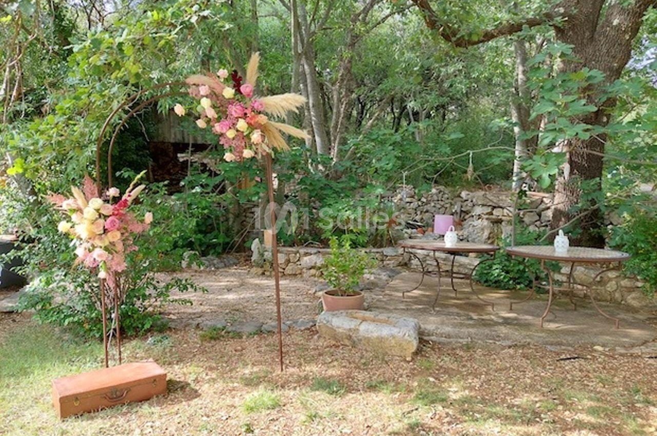 Arche florale décorée de fleurs pastel dans un jardin boisé, avec tables et pierres en arrière-plan.
