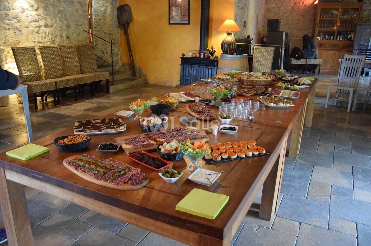 Grande table en bois garnie de plats variés, incluant charcuterie, légumes, desserts et apéritifs, dans une salle rustique.
