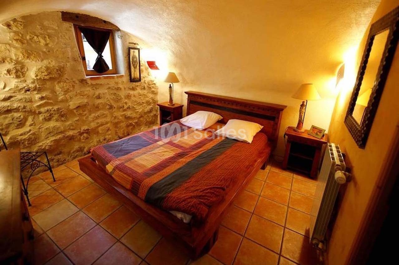 Chambre avec murs en pierre, lit double avec couvre-lit coloré, lampes de chevet et sol en carrelage.