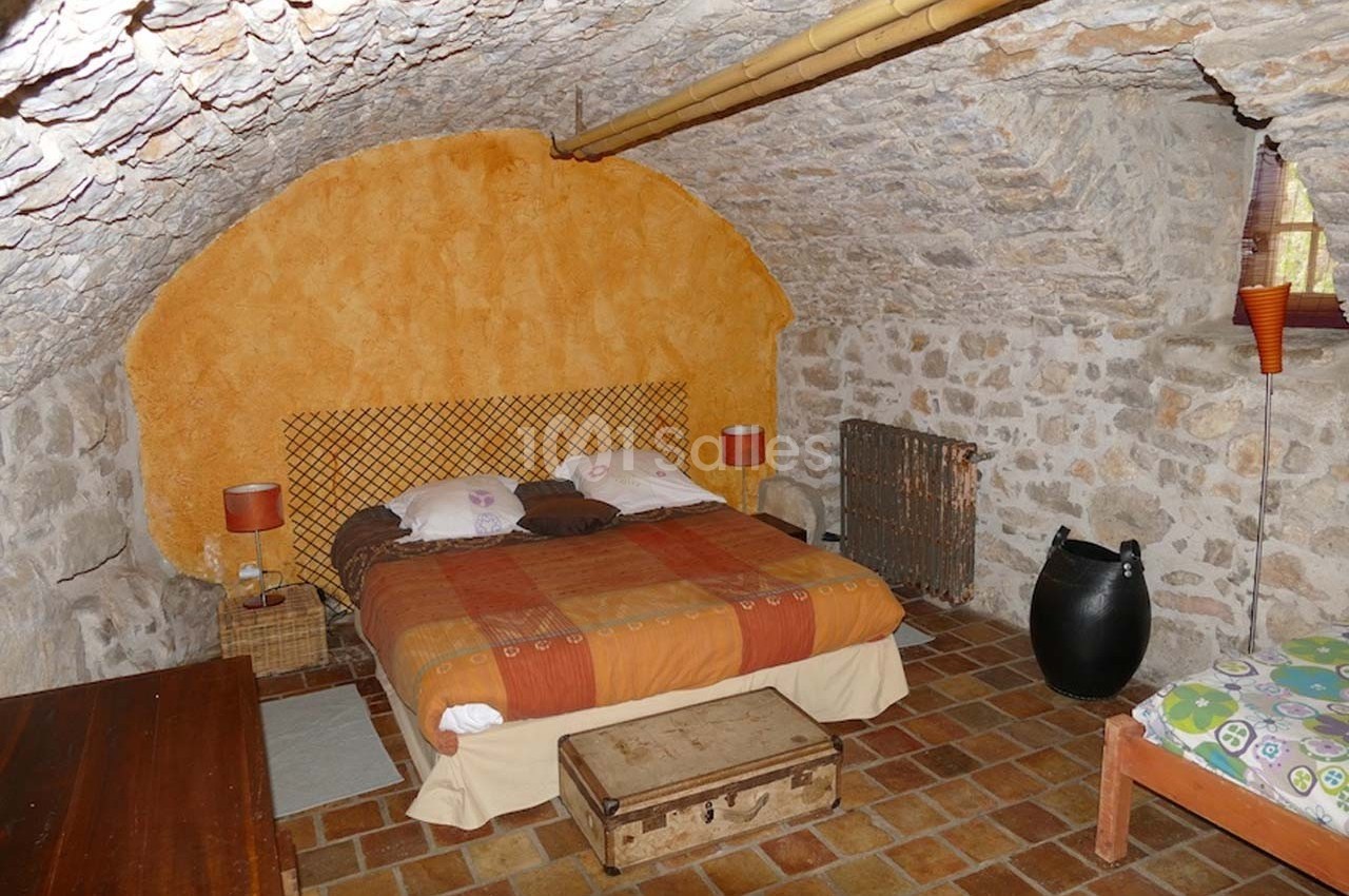 Chambre rustique avec murs en pierre, lit double, lampe de chevet, coffre en bois et sol en carreaux de terre cuite.