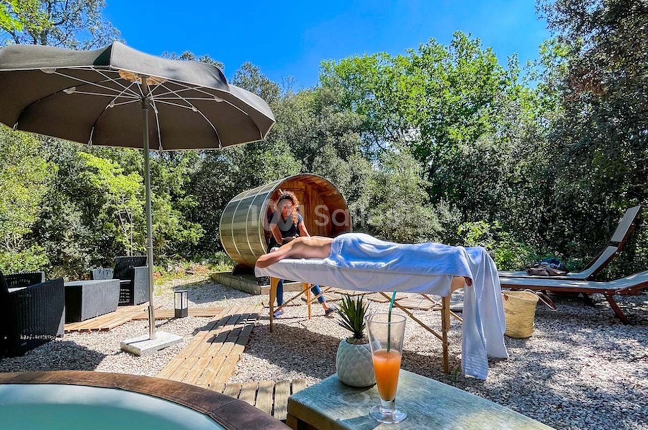 Une personne reçoit un massage en extérieur près d'un sauna en bois, entourée de végétation et de mobilier de jardin.