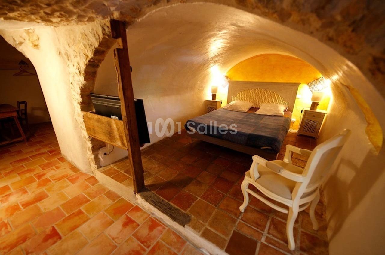 Chambre voûtée avec lit double, fauteuil blanc, sol en terre cuite et éclairage chaleureux.